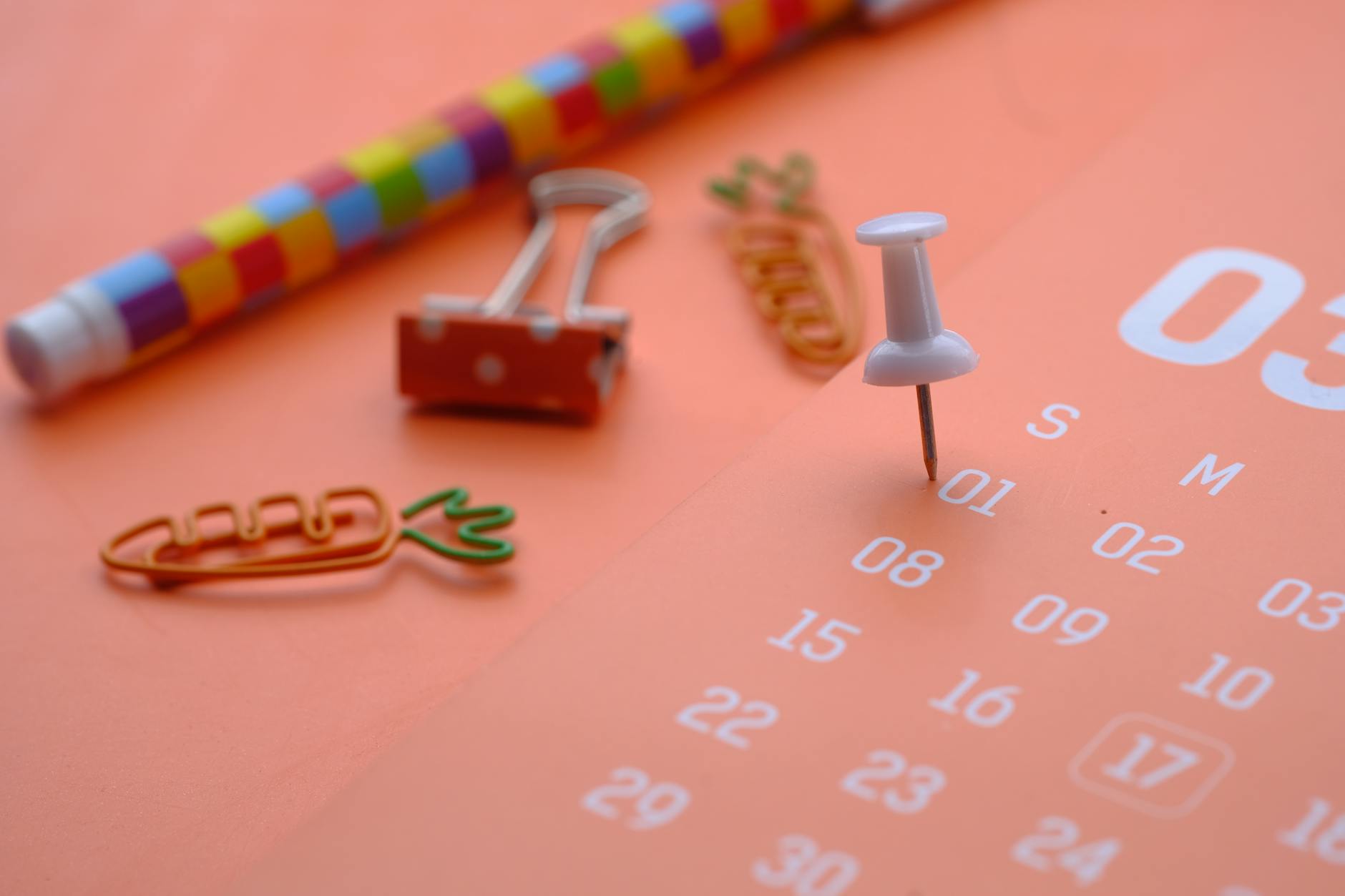 Colorful stationery items including a rainbow pen, paper clips shaped like carrots, and a white pushpin placed on a calendar page.